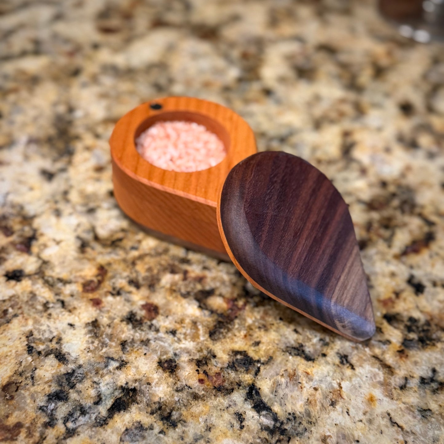 Handmade hardwood salt cellar with fitted lid, shown open on a granite countertop and filled with pink salt. Crafted from contrasting solid woods with a smooth, hand-finished surface.