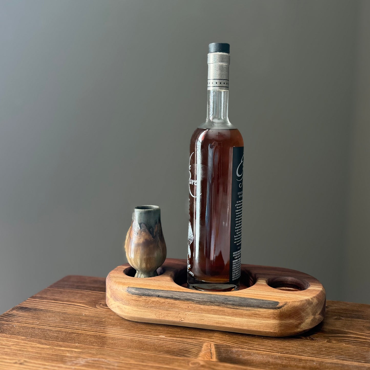 Solid hardwood whiskey bottle and glass display tray holding a bourbon bottle and rustic wooden tasting glass, photographed on a wood tabletop.