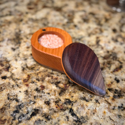 Handmade hardwood salt cellar with fitted lid, shown open on a granite countertop and filled with pink salt. Crafted from contrasting solid woods with a smooth, hand-finished surface.