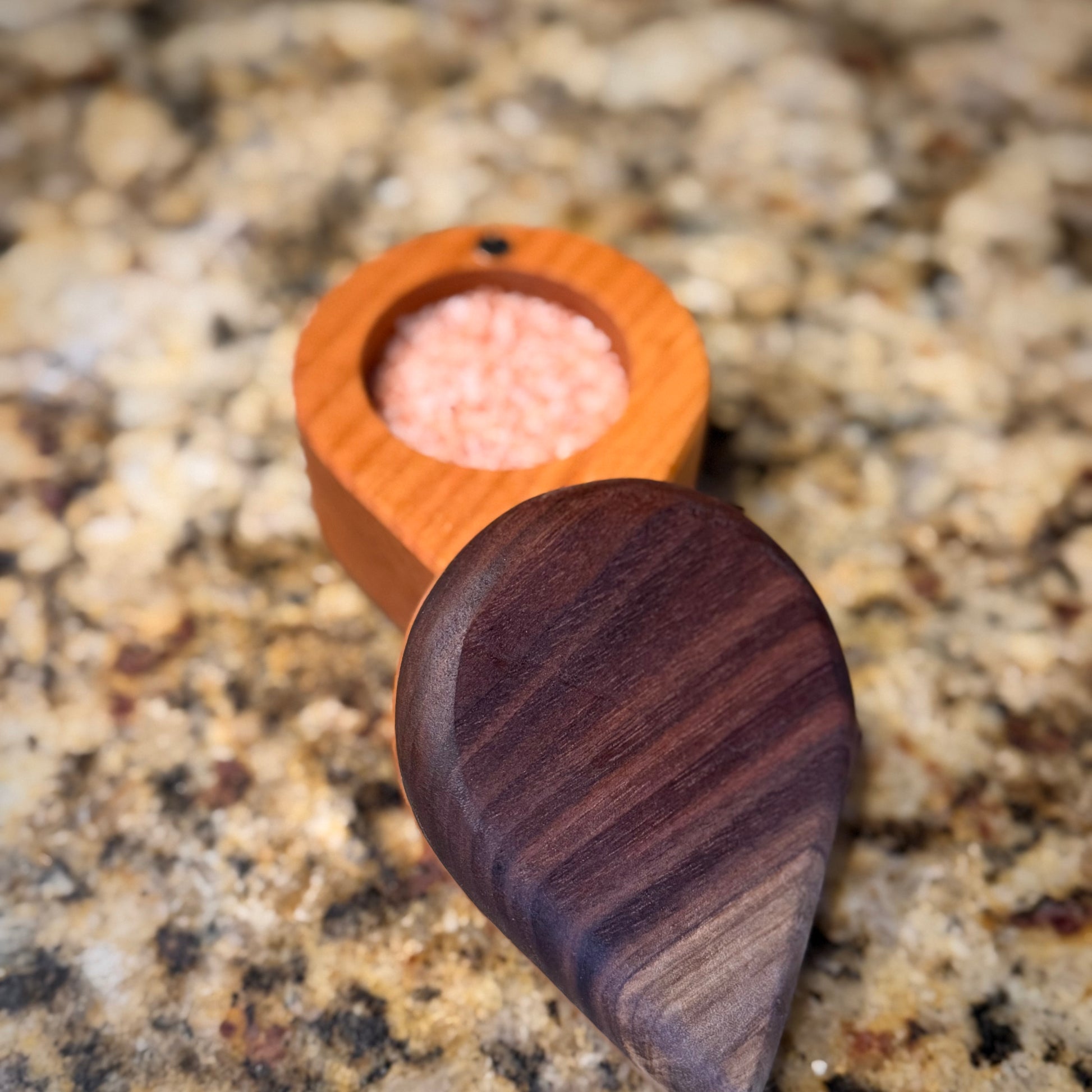 Handmade hardwood salt cellar with fitted lid, shown open on a granite countertop and filled with pink salt. Crafted from contrasting solid woods with a smooth, hand-finished surface.